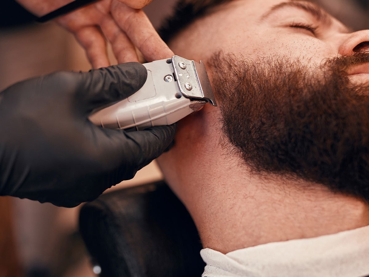 A man lying back in a barber chair with a barber using an electric razor to trim his beard. 