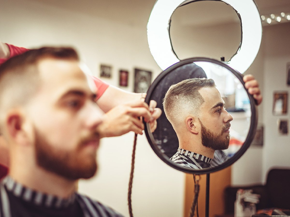 image of man in barber chair with male barber holding a mirror to the side so client can see side profile of hairline